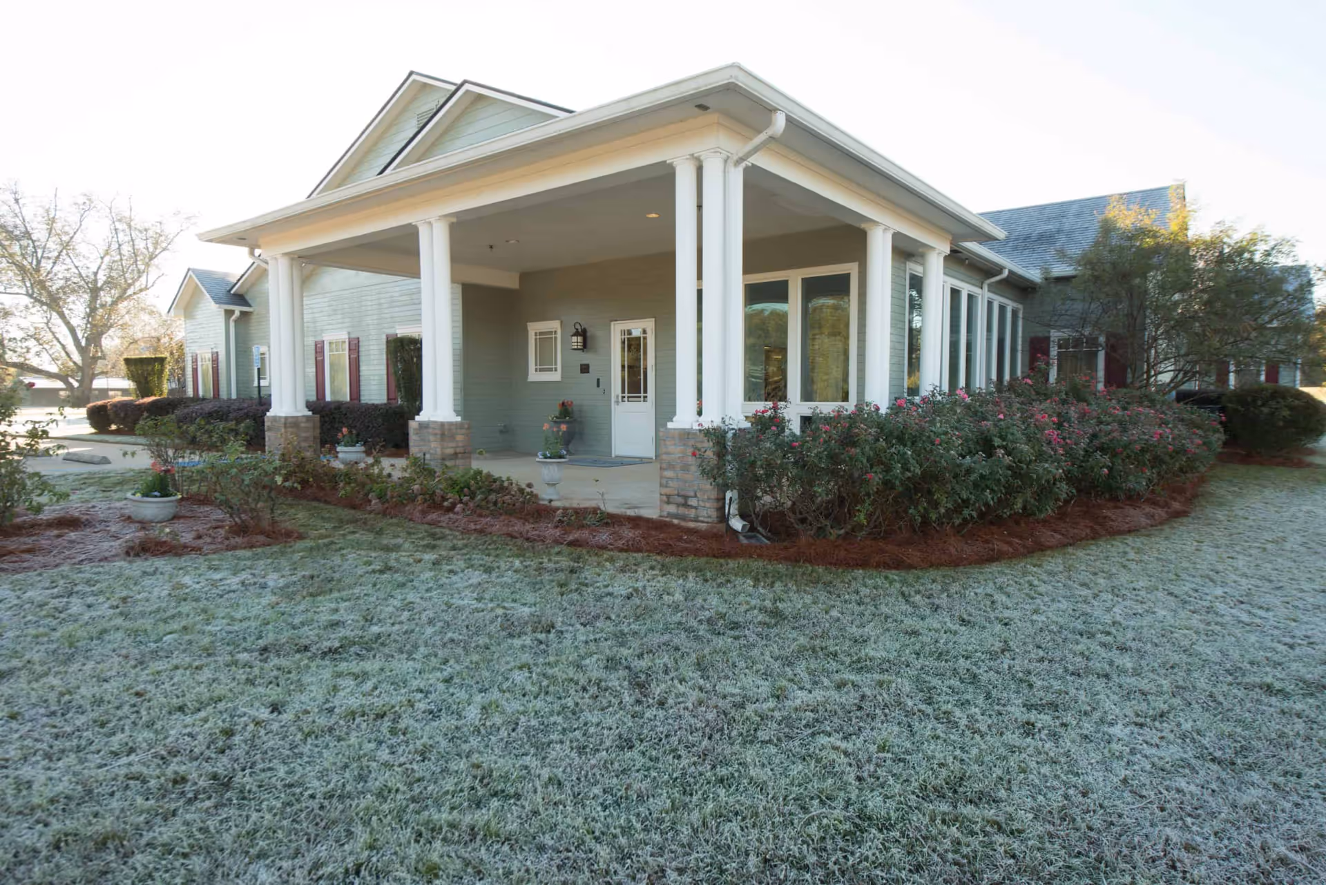 Front entrance of a single-story apartment building with a covered porch supported by white columns, shrubs and a frosty lawn in front.