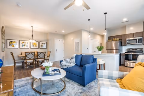 Bright and modern open-concept living space featuring a blue loveseat, a plaid armchair with a yellow pillow, a round coffee table with flowers and decor, a dining area with a wooden table and six chairs, and a kitchen with stainless steel appliances and pendant lighting.