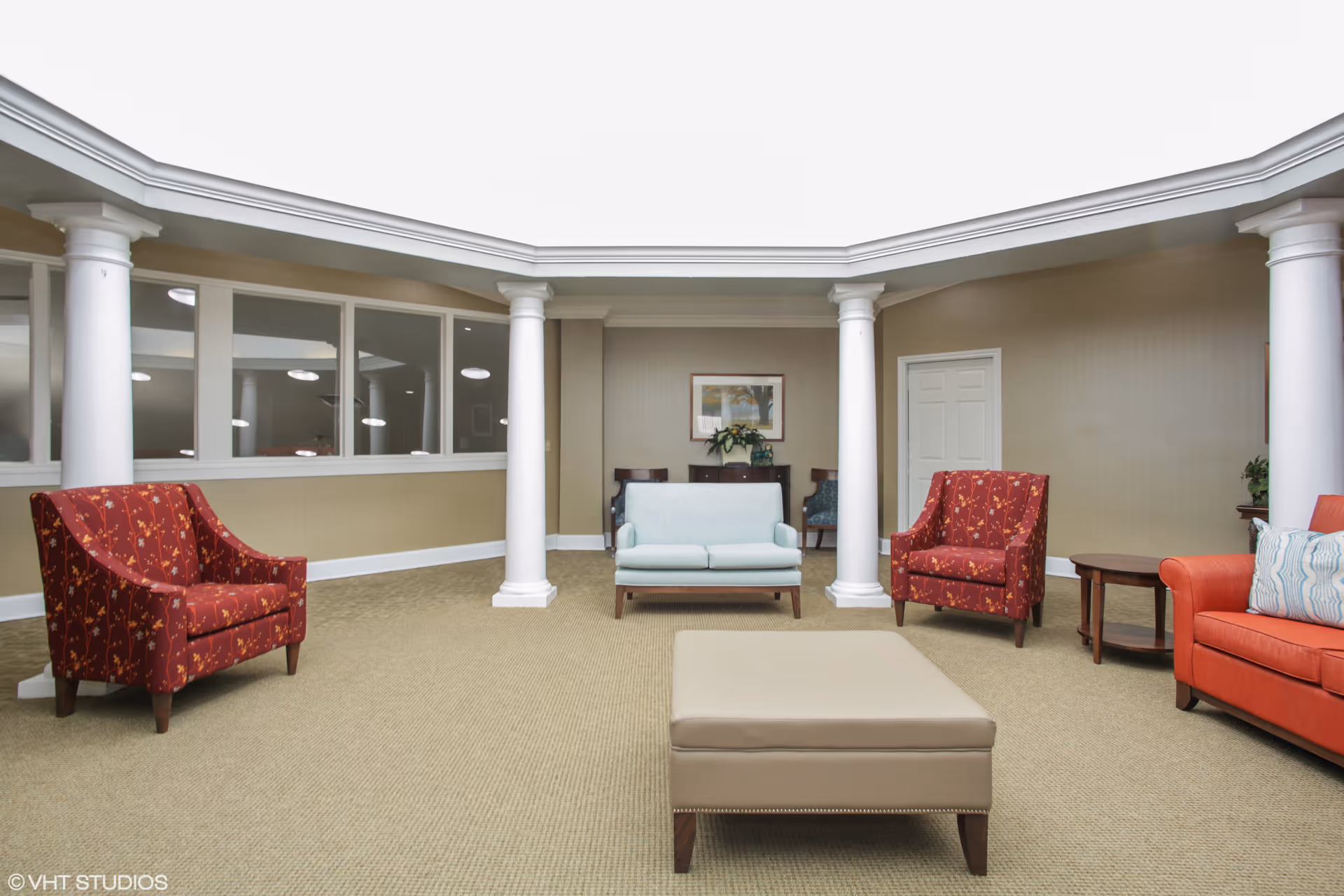 Seating area in a senior living facility lobby with columns, upholstered chairs, a couch and a central ottoman.