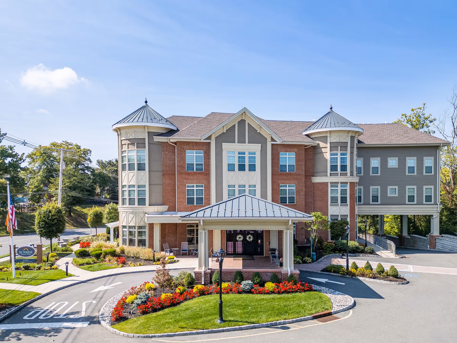 Front exterior view of Sunrise of Summit, a multi-story senior living facility with a covered entrance, landscaped flower beds, and a driveway with a stop sign painted on the pavement. The building features brick and siding with two turret-like structures on each side.
