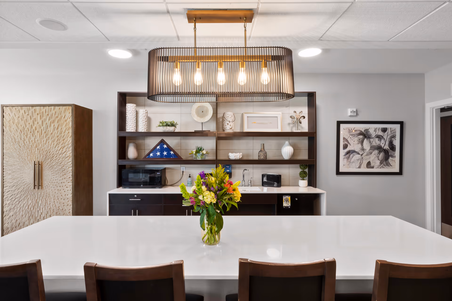 Modern kitchen area with a large white island countertop and four wooden chairs. A vase with colorful flowers is placed on the island. Behind the island, there are dark wooden shelves with decorative items including a folded American flag in a display case, vases, and plants. A microwave and toaster are on the countertop below the shelves. A large, rectangular pendant light with five bulbs hangs from the ceiling above the island. The walls are light gray with a framed abstract artwork on the right side.