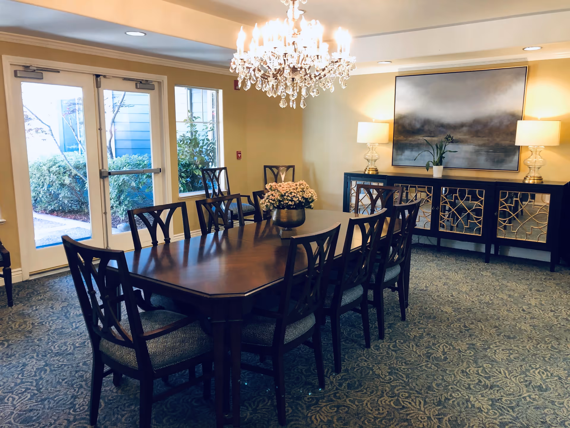 A formal dining room with a long wooden table surrounded by eight chairs. A chandelier hangs above the table, and a floral centerpiece is placed in the middle. The room has patterned carpet flooring, two large windows, and a glass door leading outside. Against the far wall is a dark wooden sideboard with mirrored cabinet doors, two table lamps, and a large framed painting above it.