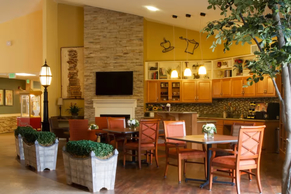 Open common dining area with round tables and chairs, a kitchenette, stone fireplace with mounted TV, and decorative plants.
