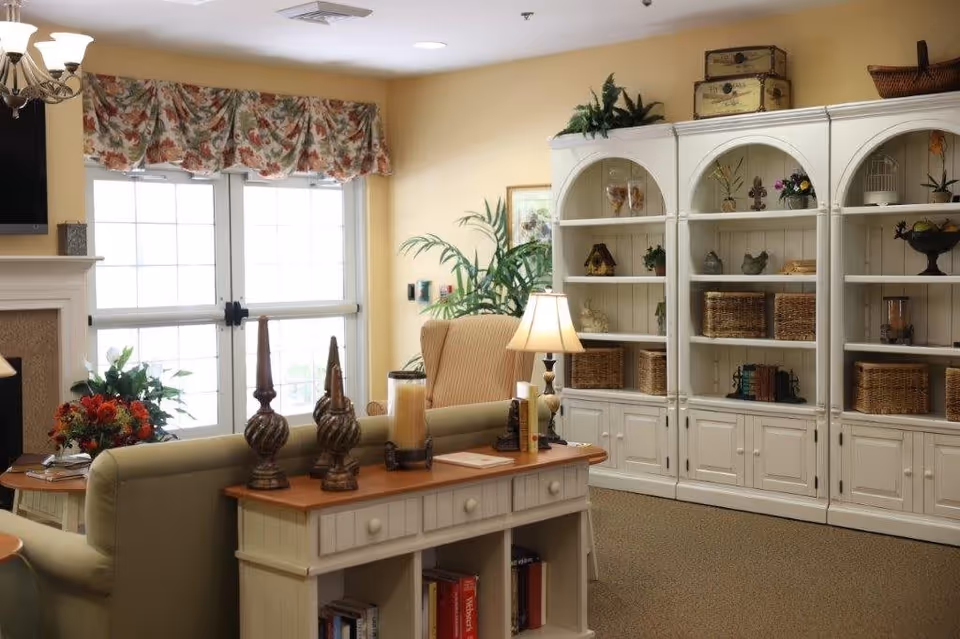 A cozy living room area with a beige sofa and armchair, a wooden console table with decorative items and books, a white built-in bookshelf filled with baskets and decor, a table lamp, a floral valance over double glass doors, and a chandelier hanging from the ceiling.