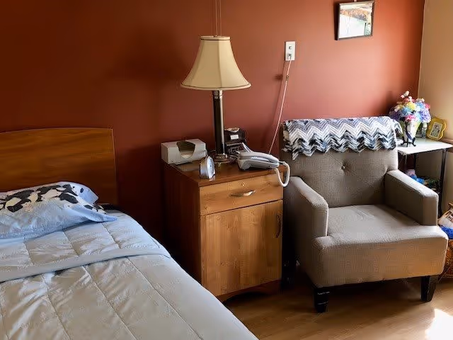 A cozy assisted living bedroom with a single bed on the left, a wooden nightstand with a lamp, telephone, and other small items in the center, and a cushioned armchair with a crocheted blanket draped over the back on the right. There is a small table with a vase of flowers and framed pictures against a reddish-brown wall.