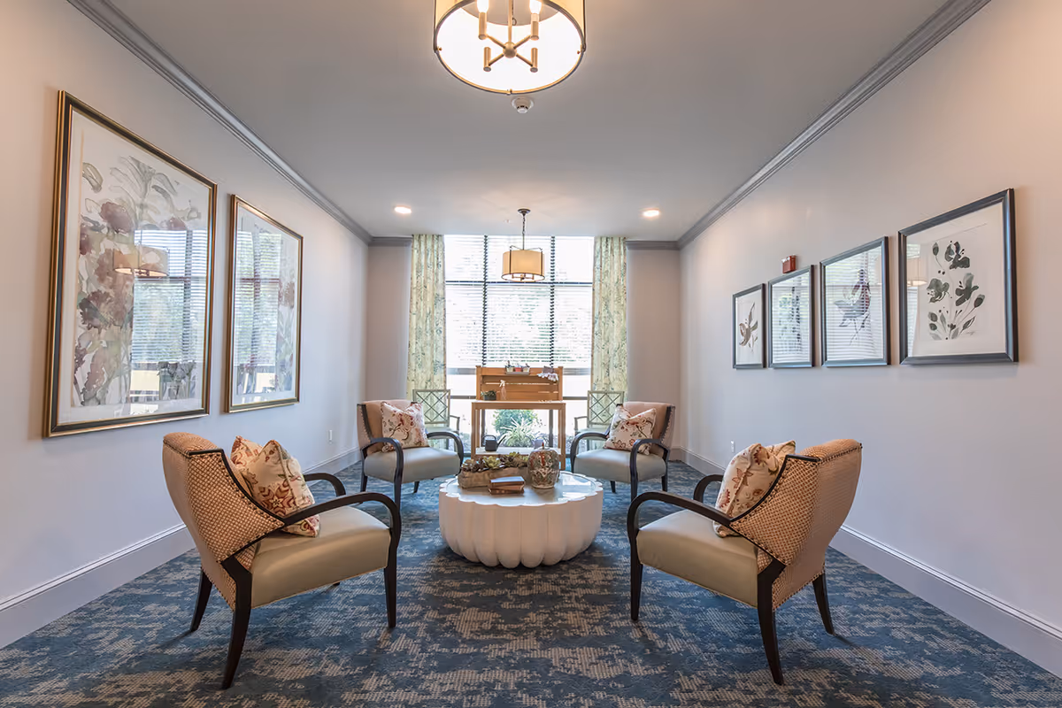 A bright and cozy sitting area with four cushioned armchairs arranged around a white round coffee table. The room has large windows with patterned curtains allowing natural light to fill the space. The walls are decorated with framed botanical artwork, and the floor is covered with a patterned carpet. Two ceiling light fixtures provide additional lighting.
