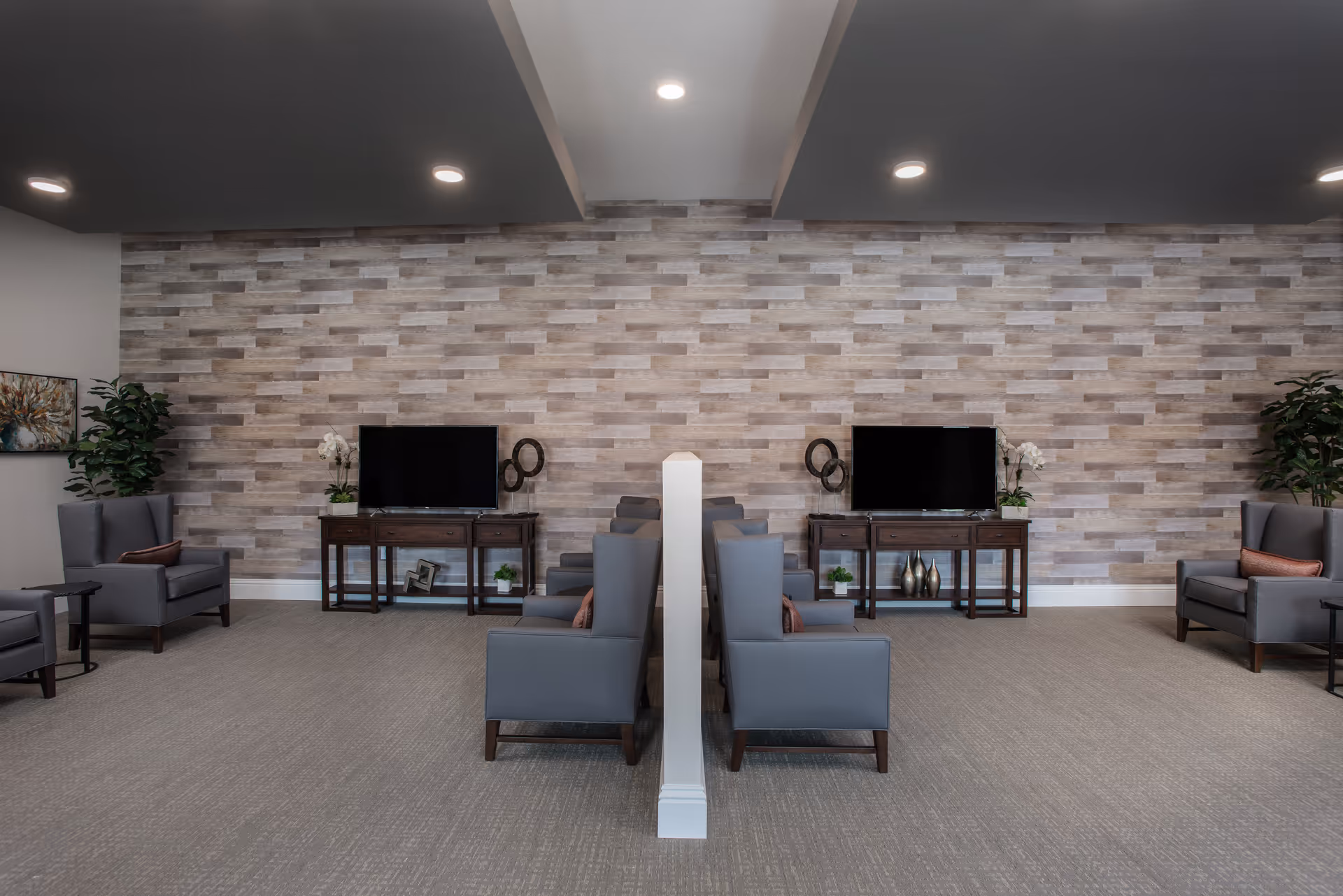 A modern lounge area with two symmetrical seating sections separated by a partial wall. Each section has gray armchairs, a wooden console table with a flat-screen TV, decorative plants, and wall art. The back wall features a textured wood panel design, and the ceiling has recessed lighting.