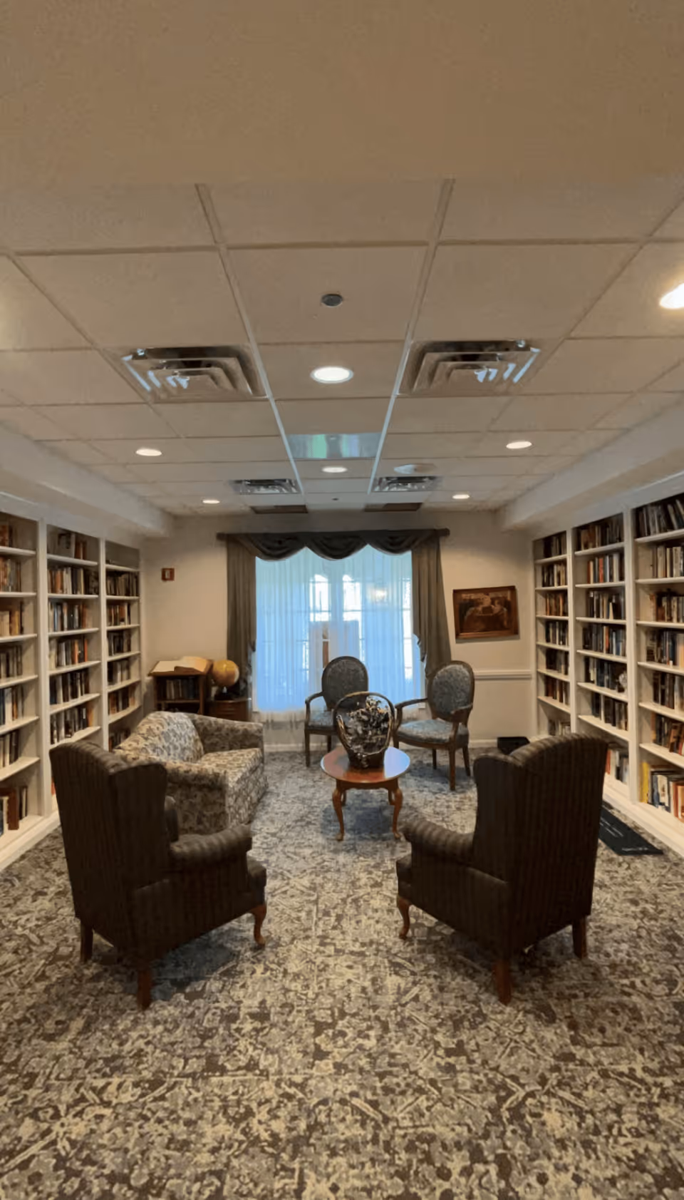 A cozy library room with bookshelves lining both walls filled with books. The room features a patterned carpet, two dark upholstered armchairs facing a small wooden coffee table with a decorative vase, a patterned sofa, and two additional chairs near a window with sheer curtains and drapes. The ceiling has recessed lighting and air vents.