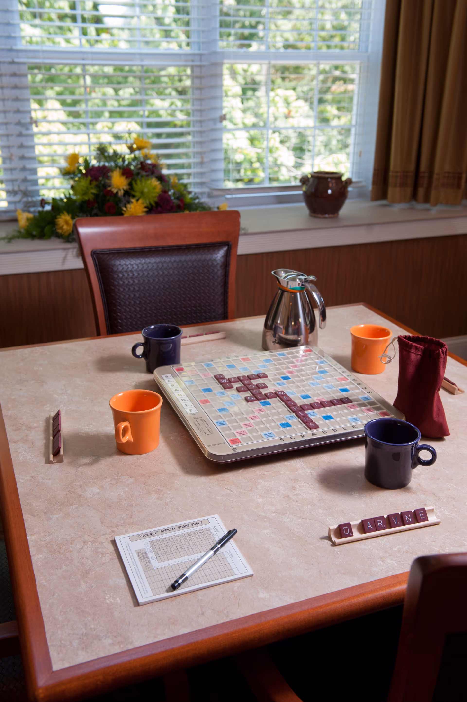 A table in a dining area set up for a Scrabble game with the board, letter racks, mugs, a score sheet, and a teapot by a window.