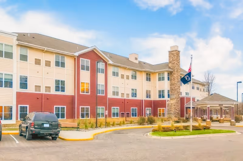 Front exterior of a three-story senior living facility with a covered entrance, flagpoles, landscaping, and parked cars.
