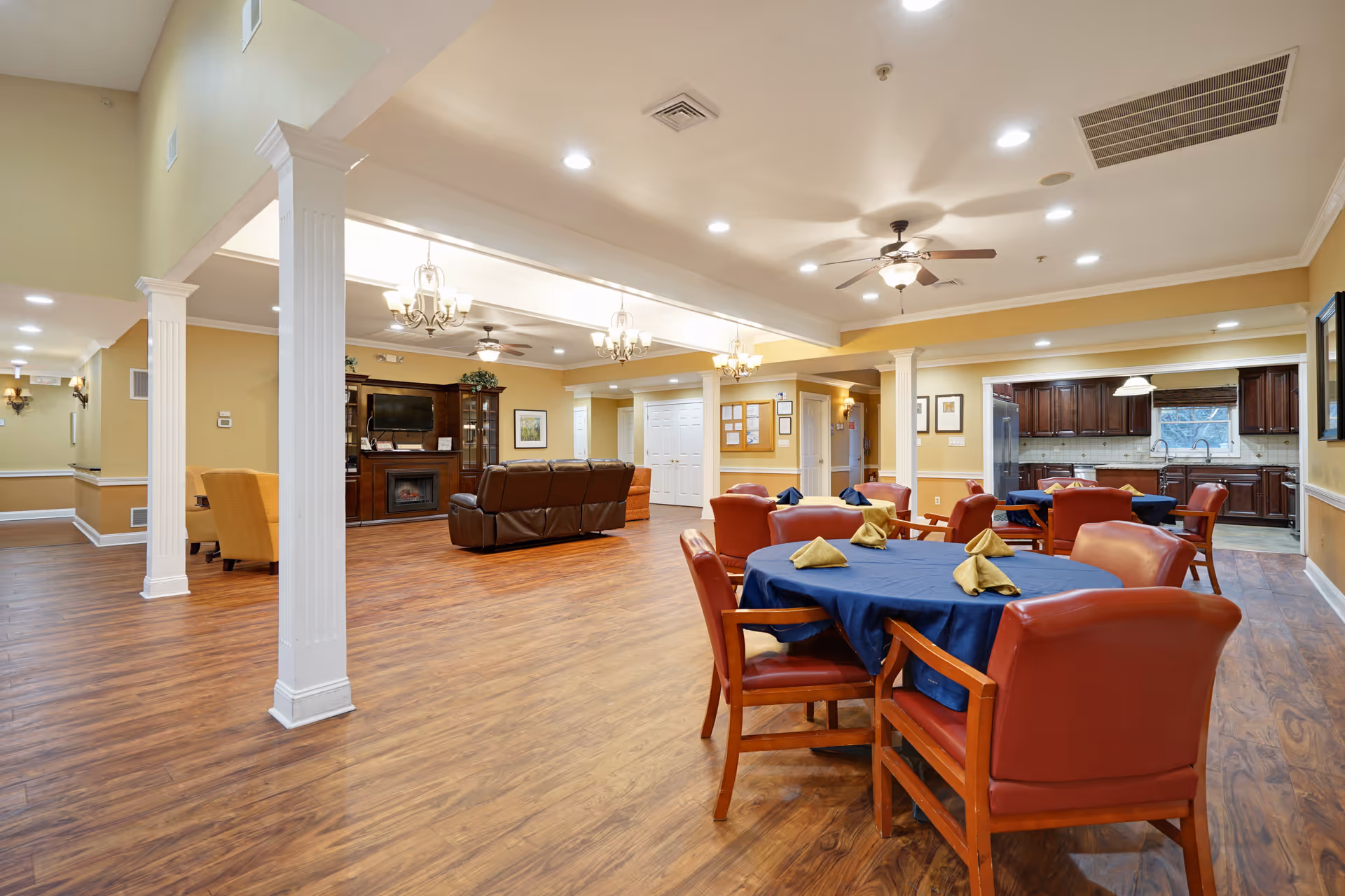 Spacious interior room with wooden flooring featuring a dining area with tables covered in blue tablecloths and red chairs, a seating area with brown leather sofas facing a TV and fireplace, and a kitchen area with dark wooden cabinets in the background. The room is well-lit with ceiling lights and chandeliers, and has beige walls with white trim and columns.