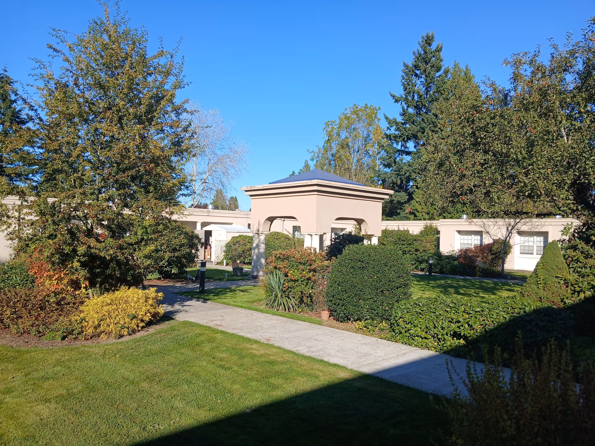 Landscaped courtyard with a paved walkway, shrubs and trees in front of a single-story beige building under a clear blue sky.