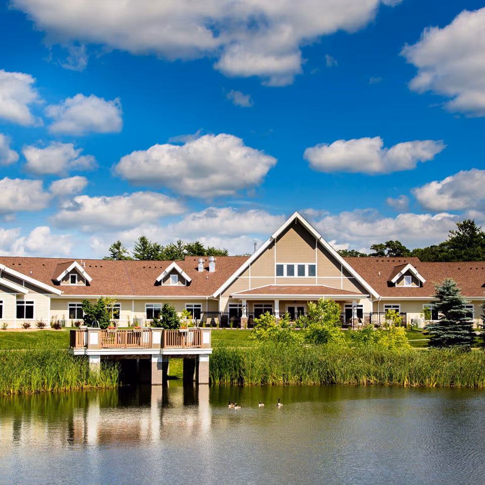 A large single-story building with a brown roof and beige siding situated behind a pond with a wooden deck extending over the water. The sky is bright blue with scattered white clouds, and there are green trees and shrubs around the building and pond.