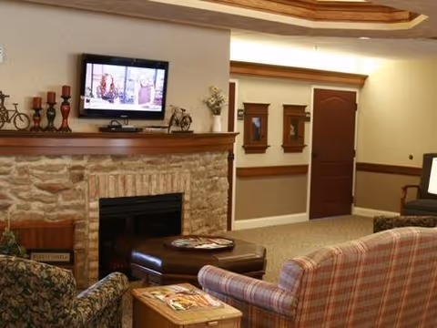 A cozy living room area with a stone fireplace and a mounted flat-screen TV above it. The room features a plaid sofa, a floral-patterned armchair, a wooden side table with magazines, and a brown leather ottoman. The walls are beige with wooden trim, and there is a closed wooden door and two decorative mirrors on the wall.