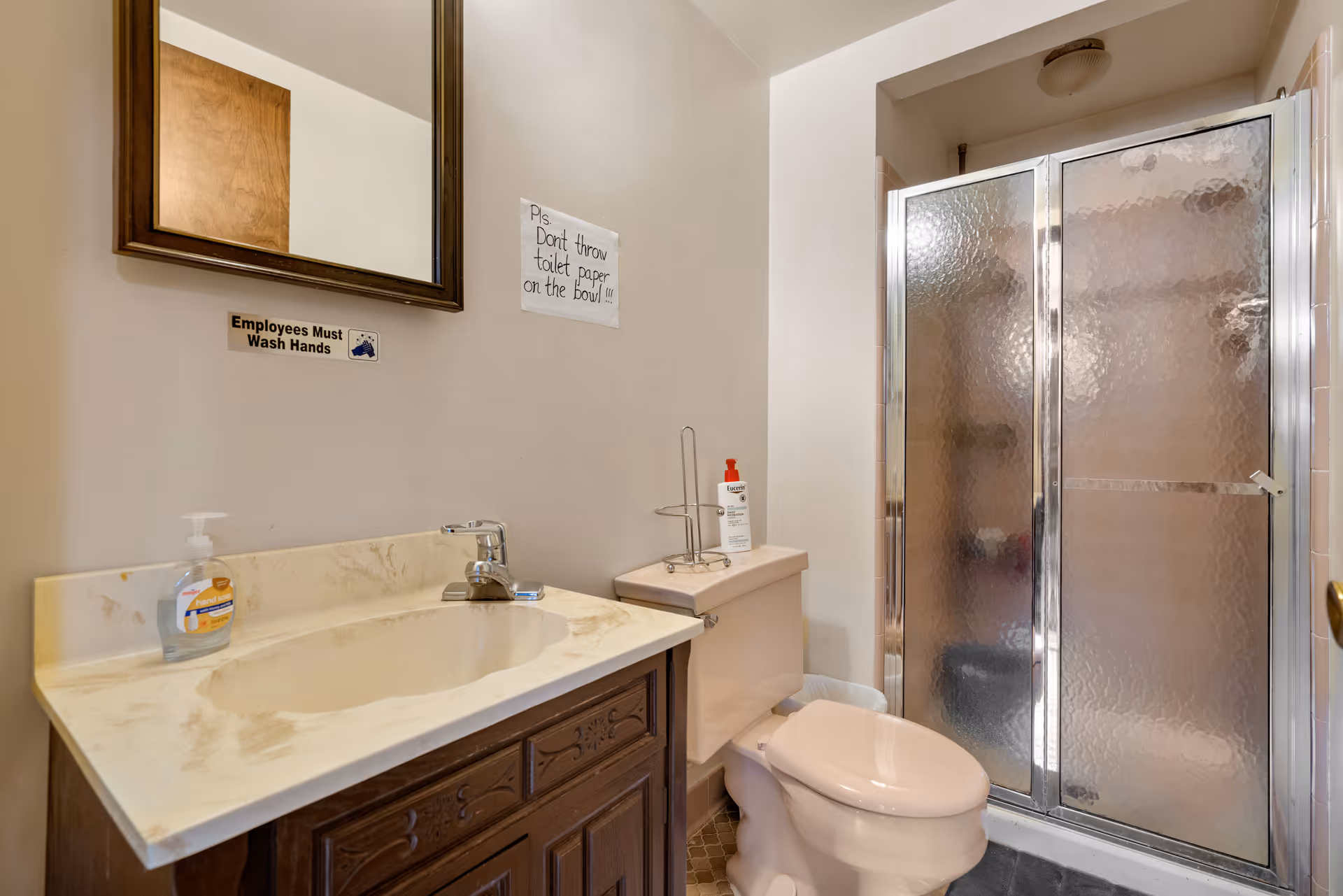 A bathroom with a beige toilet, a marble countertop sink with a wooden cabinet below, a wall-mounted mirror, and a shower with frosted glass sliding doors. There are two signs on the wall: one reads 'Employees Must Wash Hands' and the other says 'Pls Don't throw toilet paper on the bowl!!!'. A bottle of hand soap is on the sink and a bottle of hand sanitizer is on the toilet tank.