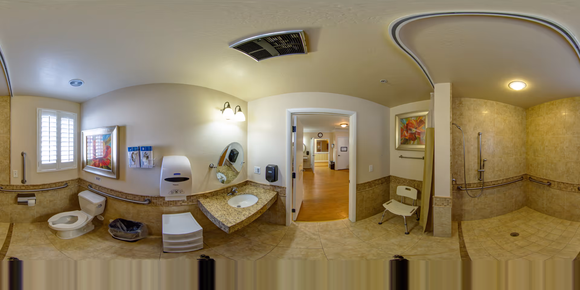 A spacious, accessible bathroom with beige tiled floors and walls. The room includes a toilet with grab bars, a sink with a granite countertop, a wall-mounted soap dispenser, a round mirror, and a paper towel dispenser. There is a shower area with grab bars and a shower chair. The bathroom has a window with white shutters, two framed floral paintings, and a door leading to a hallway with wooden flooring.