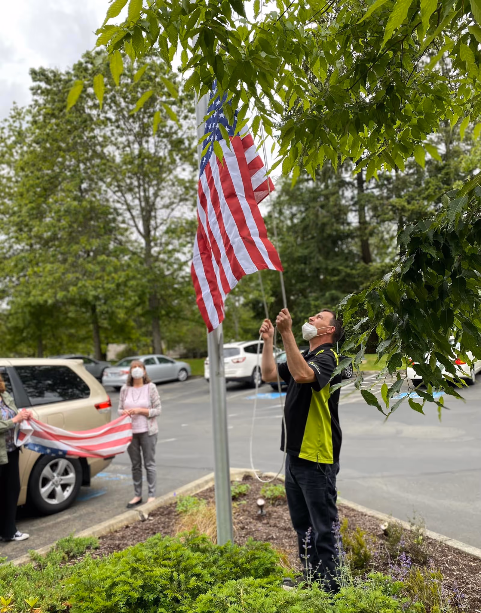 A masked man raises an American flag on a flagpole outdoors while others stand nearby holding a flag in a parking lot with cars and trees.