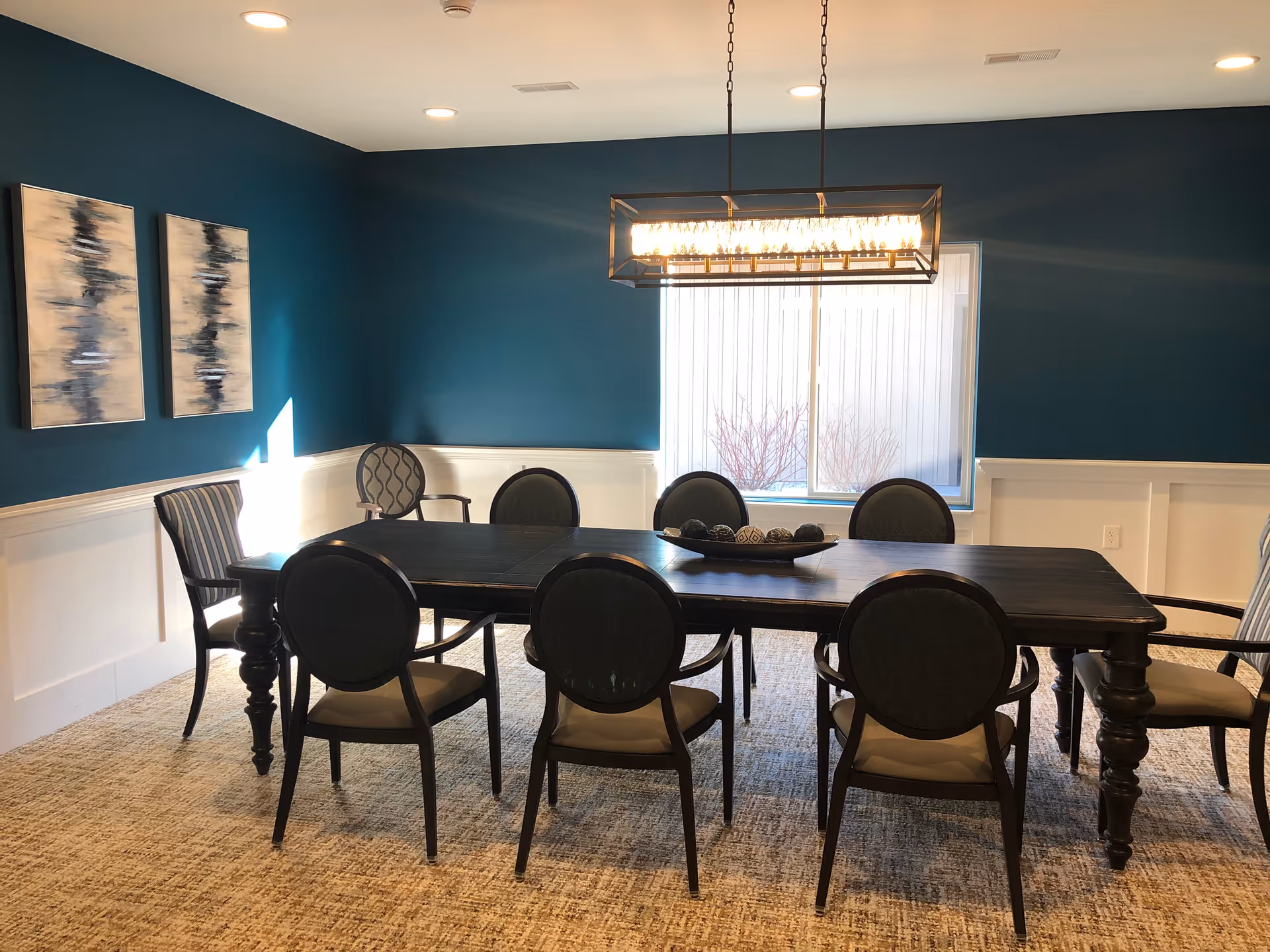 A dining room with a dark wooden rectangular table surrounded by eight chairs with cushioned seats and backs. The walls are painted teal on the upper half and white on the lower half with decorative molding. Two abstract paintings hang on the left wall. A modern rectangular chandelier hangs above the table. A window with vertical blinds is on the back wall, letting in natural light. The floor is carpeted with a textured pattern.