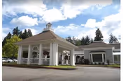 Exterior view of a senior living facility with a covered entrance featuring white columns and hanging flower baskets. The building is surrounded by trees under a partly cloudy sky.