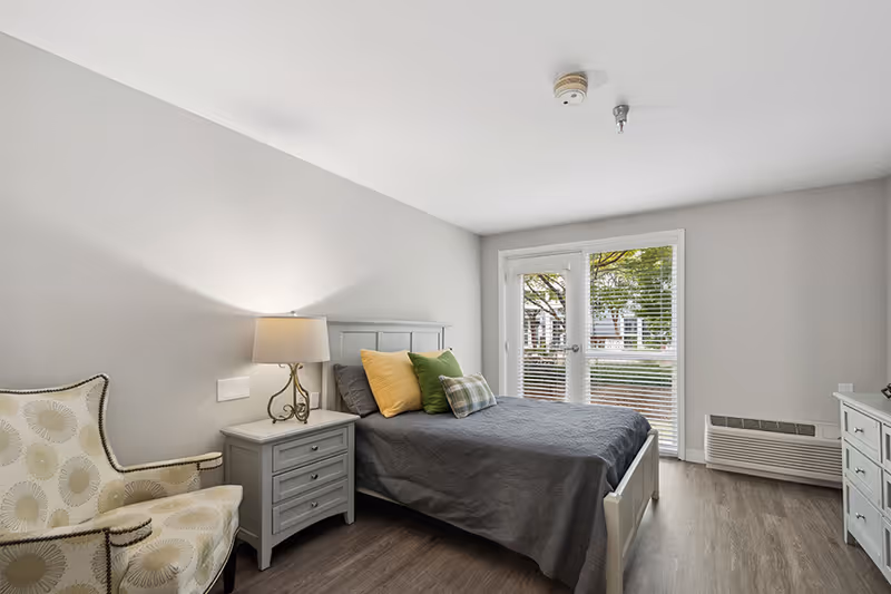 A bright bedroom with a gray bed covered with a dark gray blanket and three pillows in yellow, green, and plaid. Next to the bed is a gray nightstand with a lamp. There is a patterned armchair to the left and a dresser to the right. The room has light gray walls, wood flooring, and a glass door with blinds leading outside.
