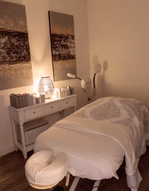 A cozy treatment room with a massage table covered in white and patterned linens. A white side table next to the table holds a glowing decorative lamp, several small bottles, and boxes. Two abstract paintings hang on the wall above the side table. The room has warm lighting and wooden flooring.