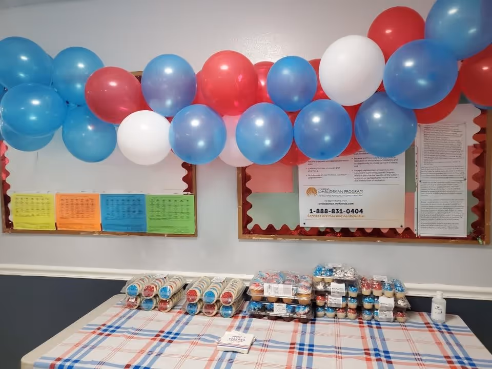 A table covered with a red, white, and blue plaid tablecloth displaying trays of cupcakes decorated in red, white, and blue. Above the table, there is a garland of red, white, and blue balloons. Behind the table, there are bulletin boards with various notices and papers pinned on them.
