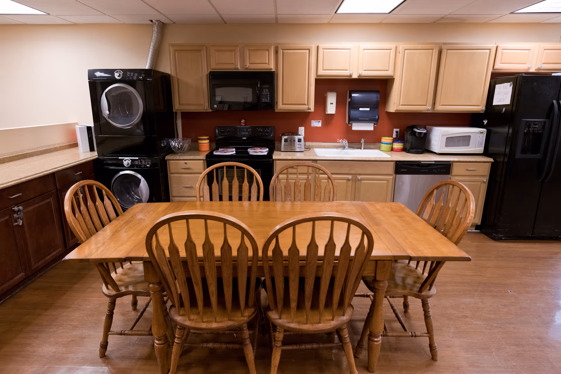 A kitchen area with wooden cabinets, a black refrigerator, microwave, dishwasher, stove, and a stacked washer and dryer. In the center, there is a wooden dining table with six matching wooden chairs. The walls are painted beige and red, and the floor is wooden.