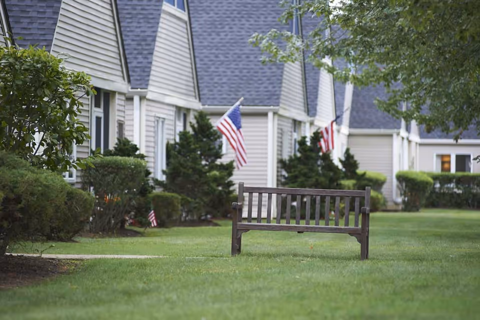A wooden bench on a well-maintained grassy lawn in front of a row of beige residential buildings with gray roofs. Several American flags are displayed along the buildings, and there are bushes and trees around the area.
