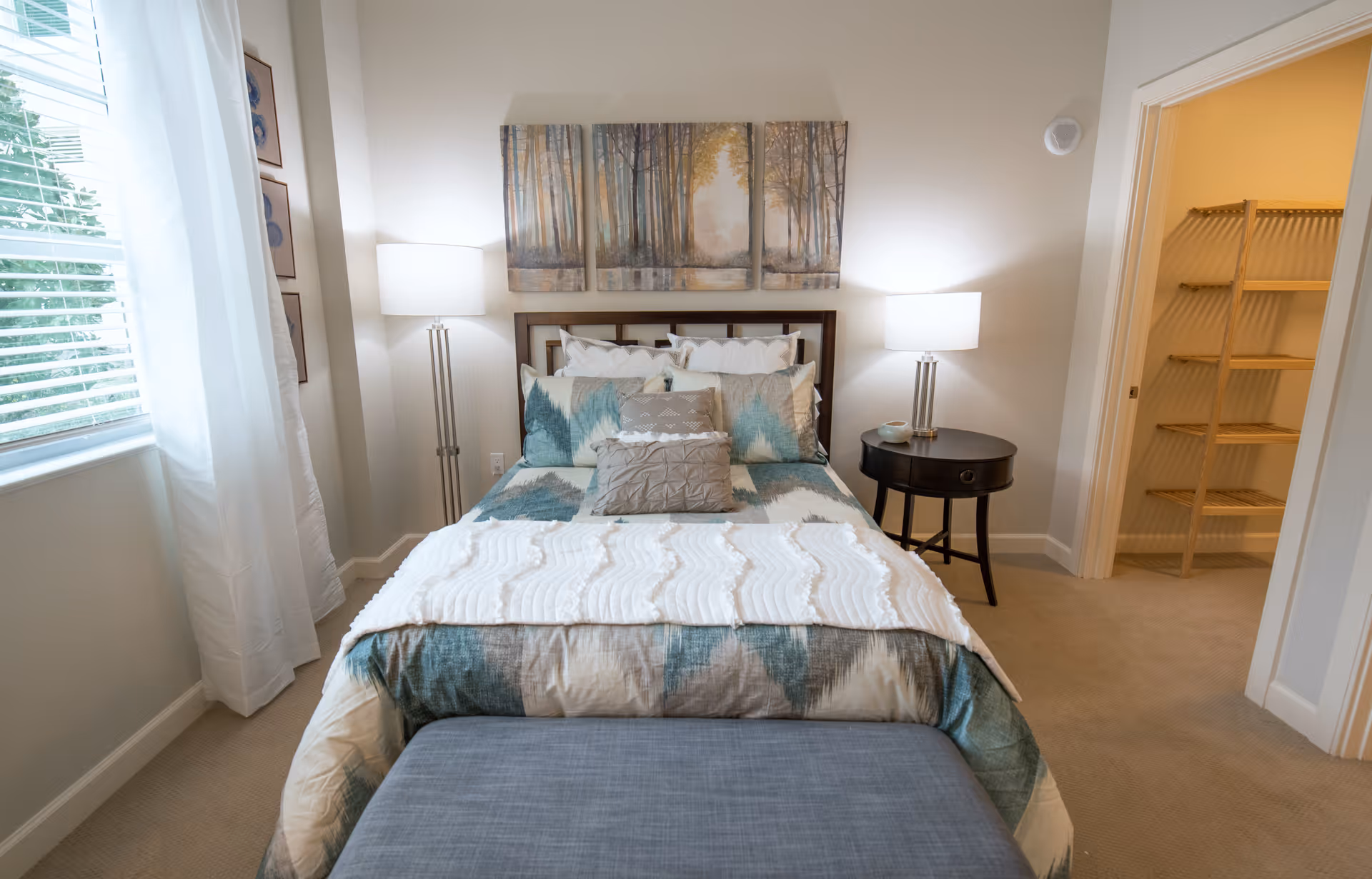 Well-lit furnished bedroom with a made bed, bedside tables and lamps, artwork above the headboard, and a walk-in closet to the right.