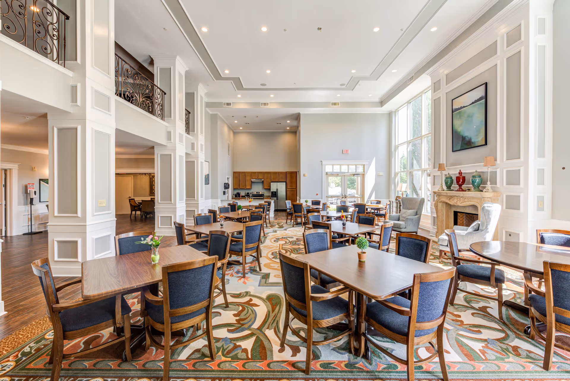 A spacious and elegant dining area in a senior living facility with multiple wooden tables and blue cushioned chairs arranged on a patterned carpet. The room features high ceilings with recessed lighting, large windows allowing natural light to fill the space, a decorative fireplace with vases and lamps on the mantel, and a kitchen area with wooden cabinets and stainless steel appliances in the background.