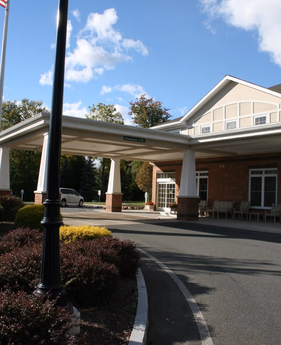 Covered porte-cochère entrance of a brick senior living building with a driveway, benches, and landscaped shrubs under a blue sky.