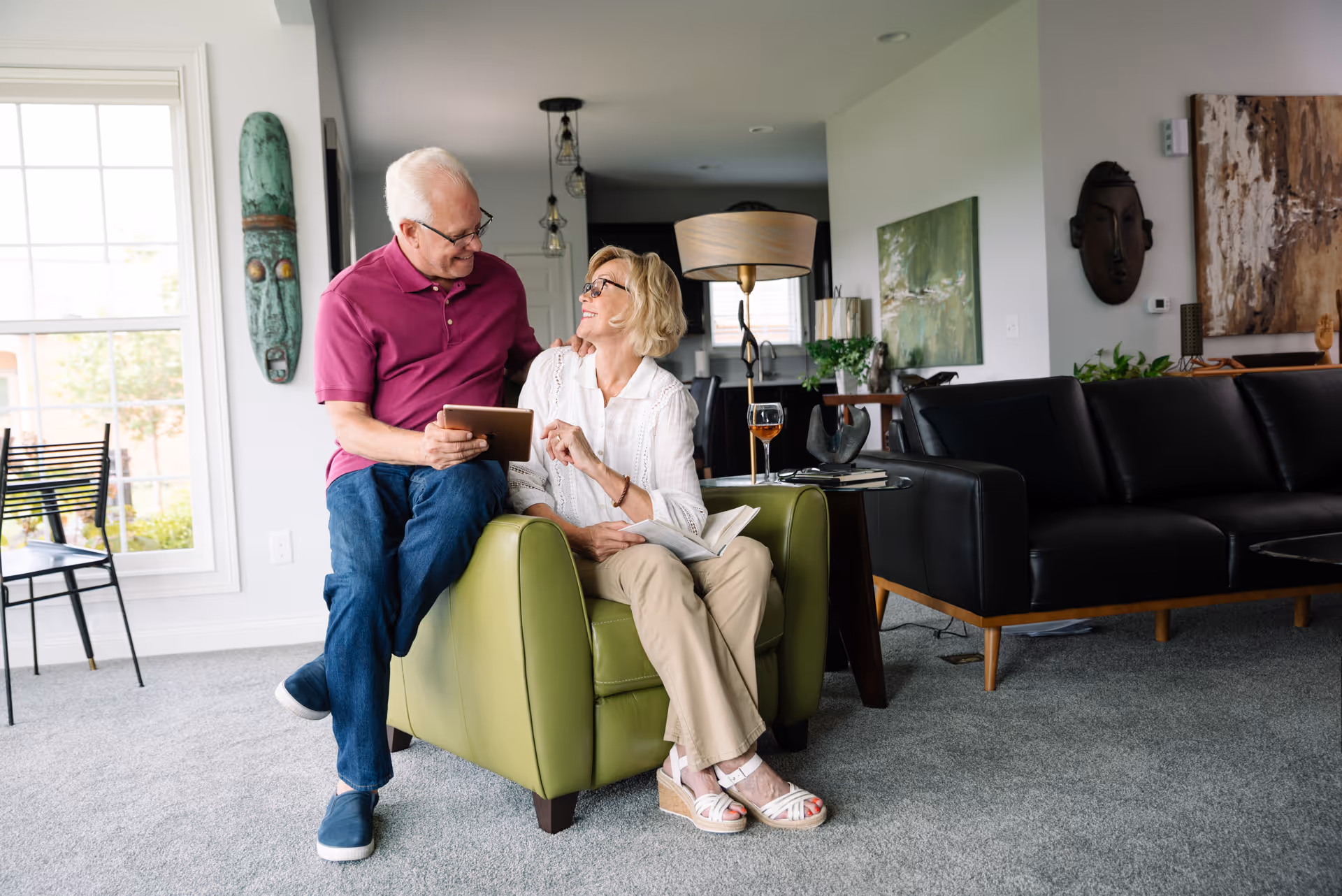 An elderly couple sitting in a modern living room. The man in a magenta polo shirt and jeans is sitting on the arm of a green armchair, holding a tablet and smiling at the woman. The woman, wearing a white blouse and beige pants, is seated in the armchair, looking up at the man with a smile. The room features a black leather sofa, a side table with a glass of wine, decorative wall art, and a large window letting in natural light.