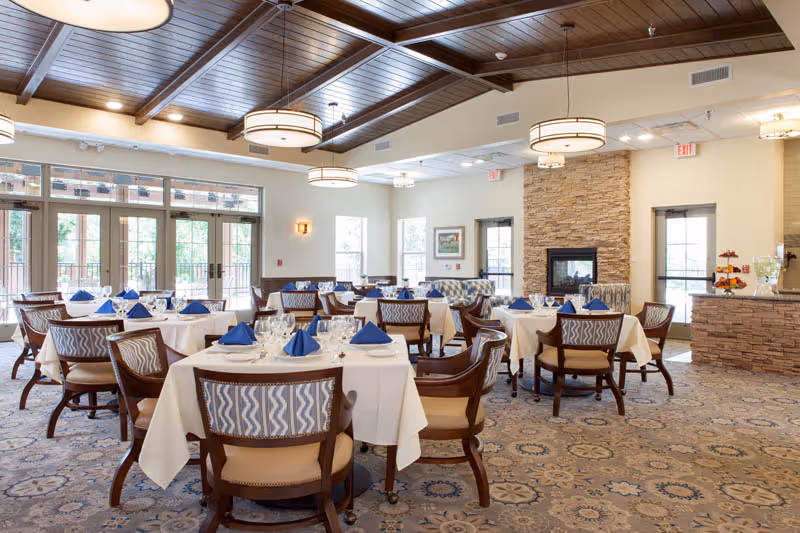 A spacious dining room with multiple round tables covered with white tablecloths and set with blue napkins, glassware, and silverware. The room features large windows and glass doors letting in natural light, a stone fireplace, patterned carpet, and wooden ceiling beams with modern circular light fixtures.