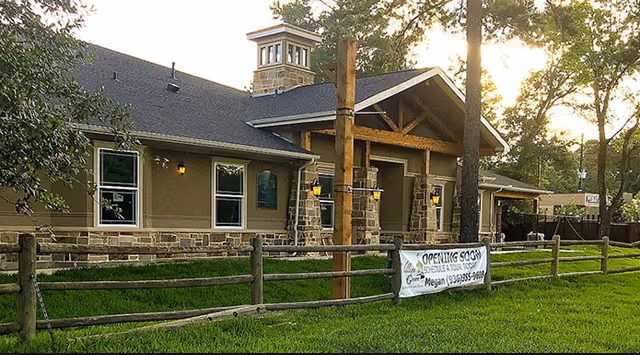 Front exterior of a single-story memory care building with stone pillars, a covered wooden entrance, and an 'Opening Soon' banner on a split-rail fence.
