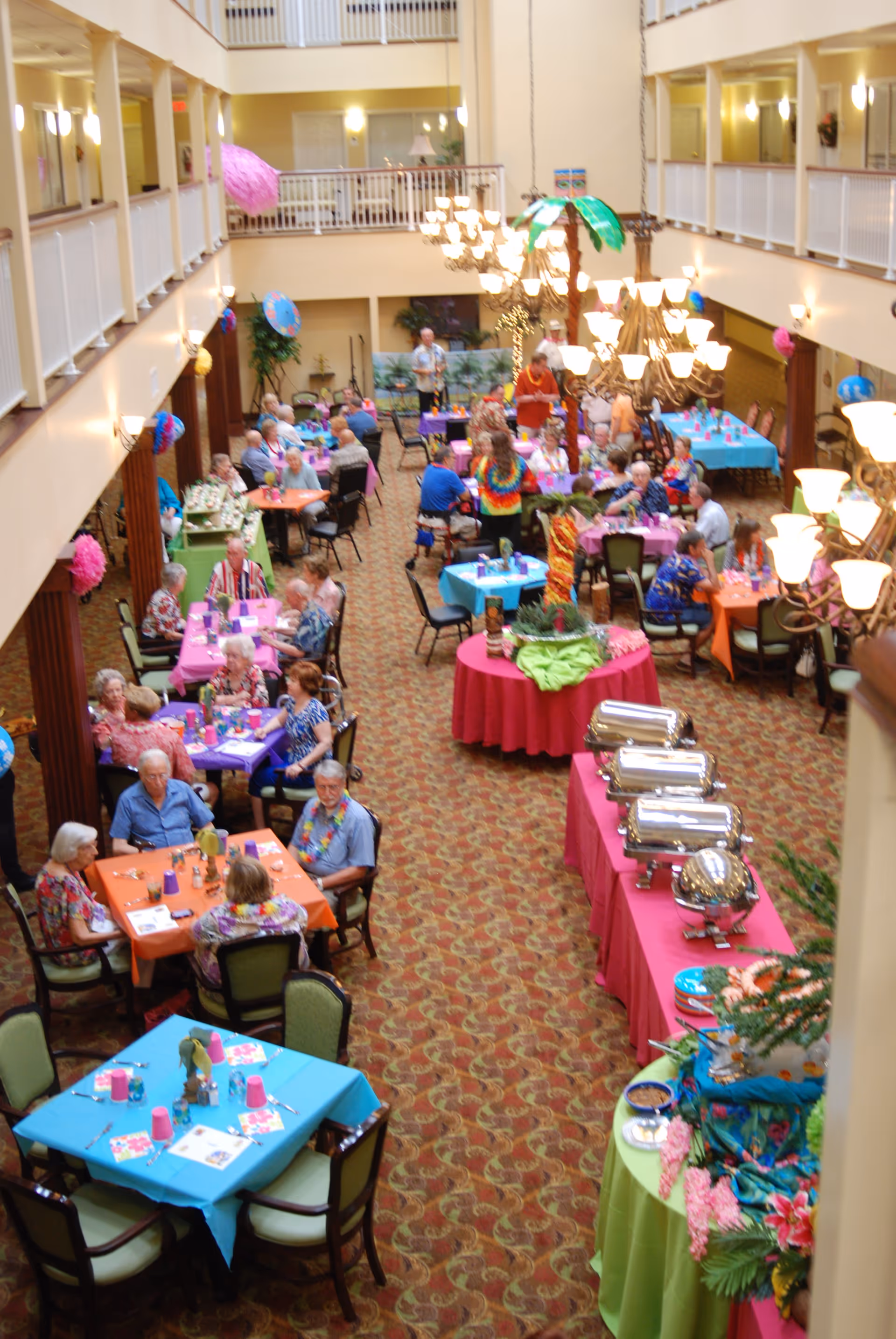A large dining area in a senior living community decorated with colorful tablecloths and tropical-themed decorations. Several elderly people are seated at tables, enjoying a meal or socializing. Buffet tables with covered dishes are set up along one side of the room. The space has high ceilings with chandeliers and a balcony overlooking the dining area.