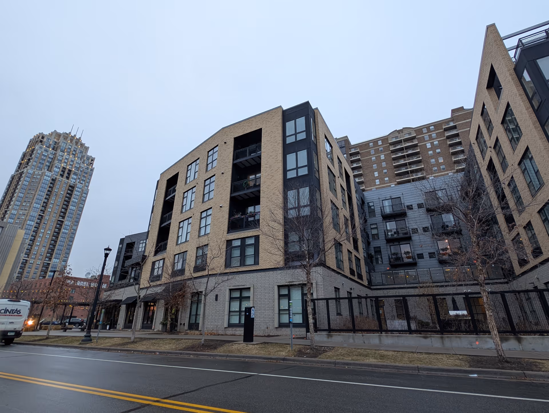 Exterior view of Abiitan Mill City, a multi-story residential building with beige and gray brick facade, large windows, and balconies. The street in front is wet, and there are leafless trees and a streetlamp along the sidewalk. A tall building is visible in the background under a cloudy sky.
