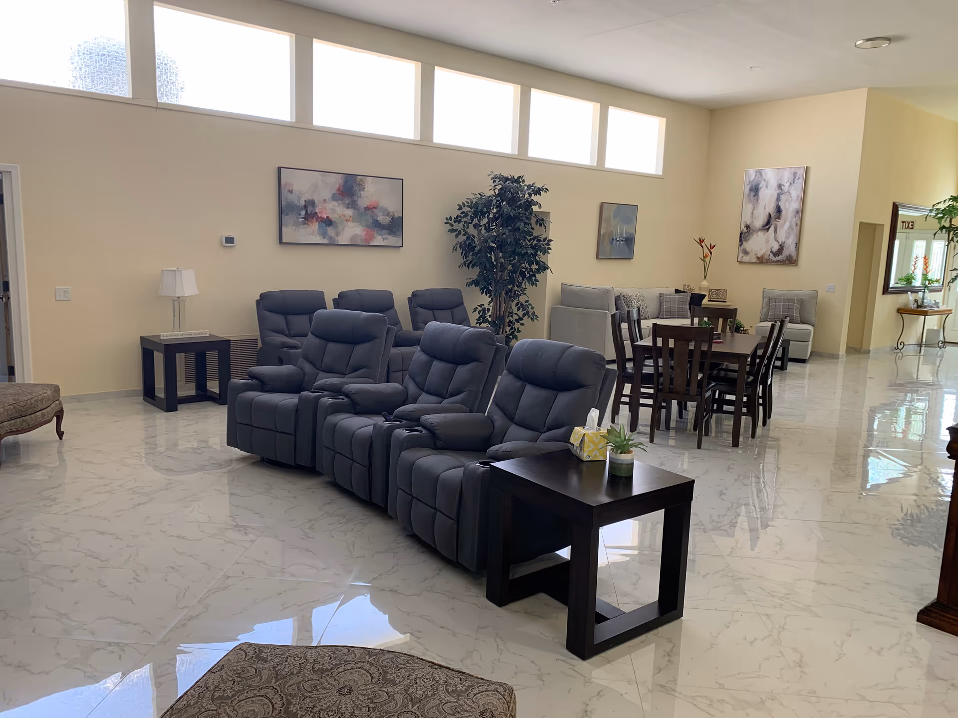 A spacious and well-lit common living area in Meadow Assisted Living Care featuring a row of four gray recliner chairs with a small dark wooden side table holding a tissue box and a small plant. Behind the recliners is a dining table with six chairs, a gray couch, and two armchairs. The room has large windows near the ceiling, light-colored walls, marble-patterned tile flooring, and several pieces of abstract wall art and plants.