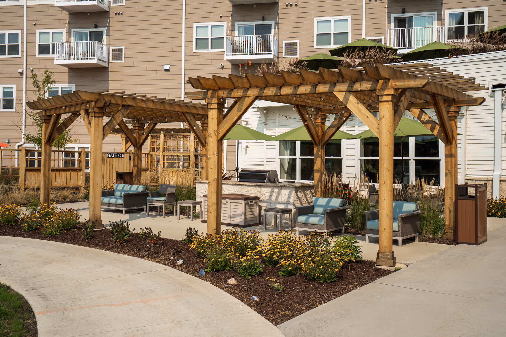 Outdoor courtyard showing wooden pergolas with cushioned seating, tables, and landscaping in front of a multi-story senior living building.