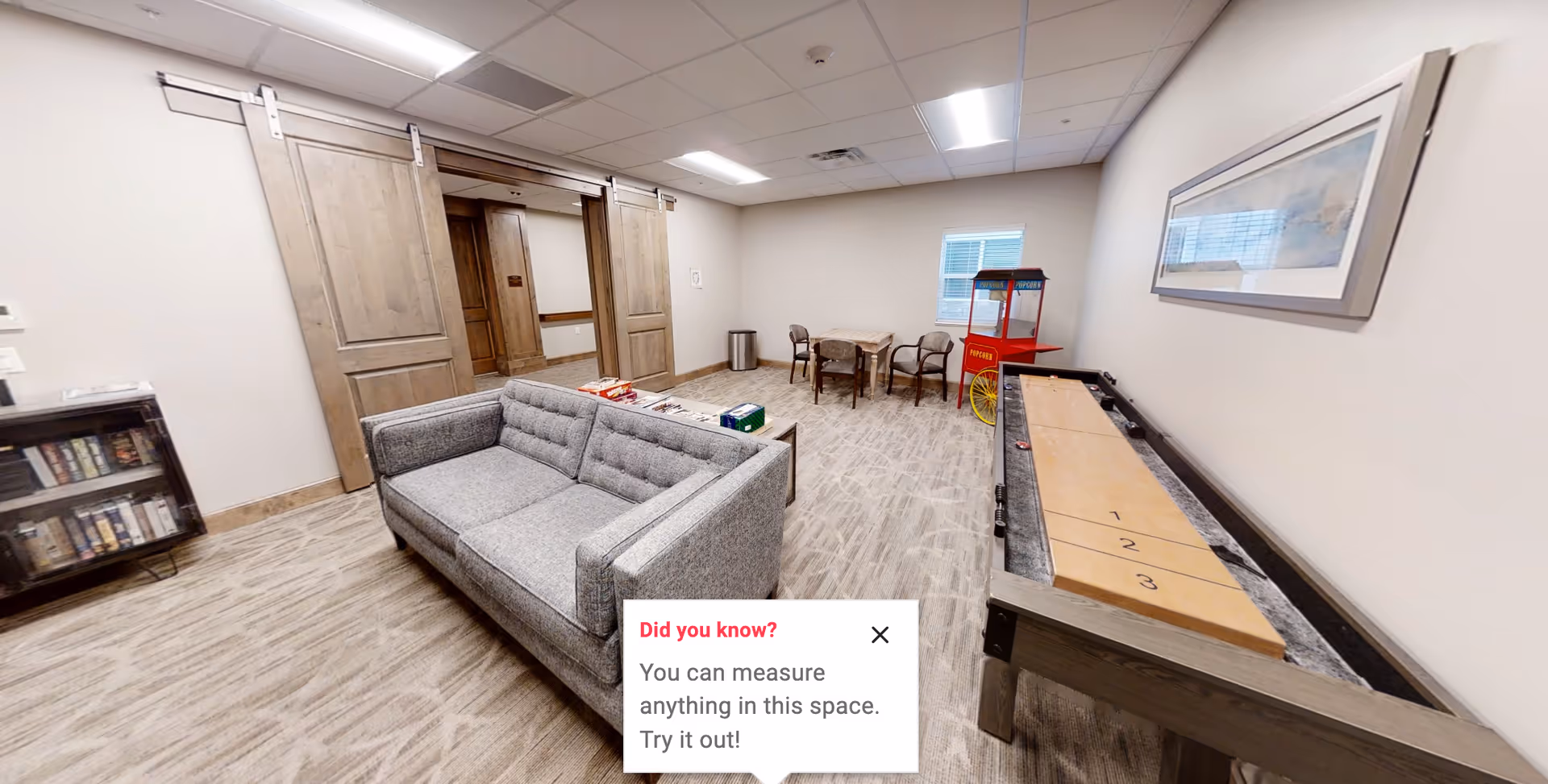 A cozy activity room with a gray couch, shuffleboard table, small dining table, and a popcorn cart.