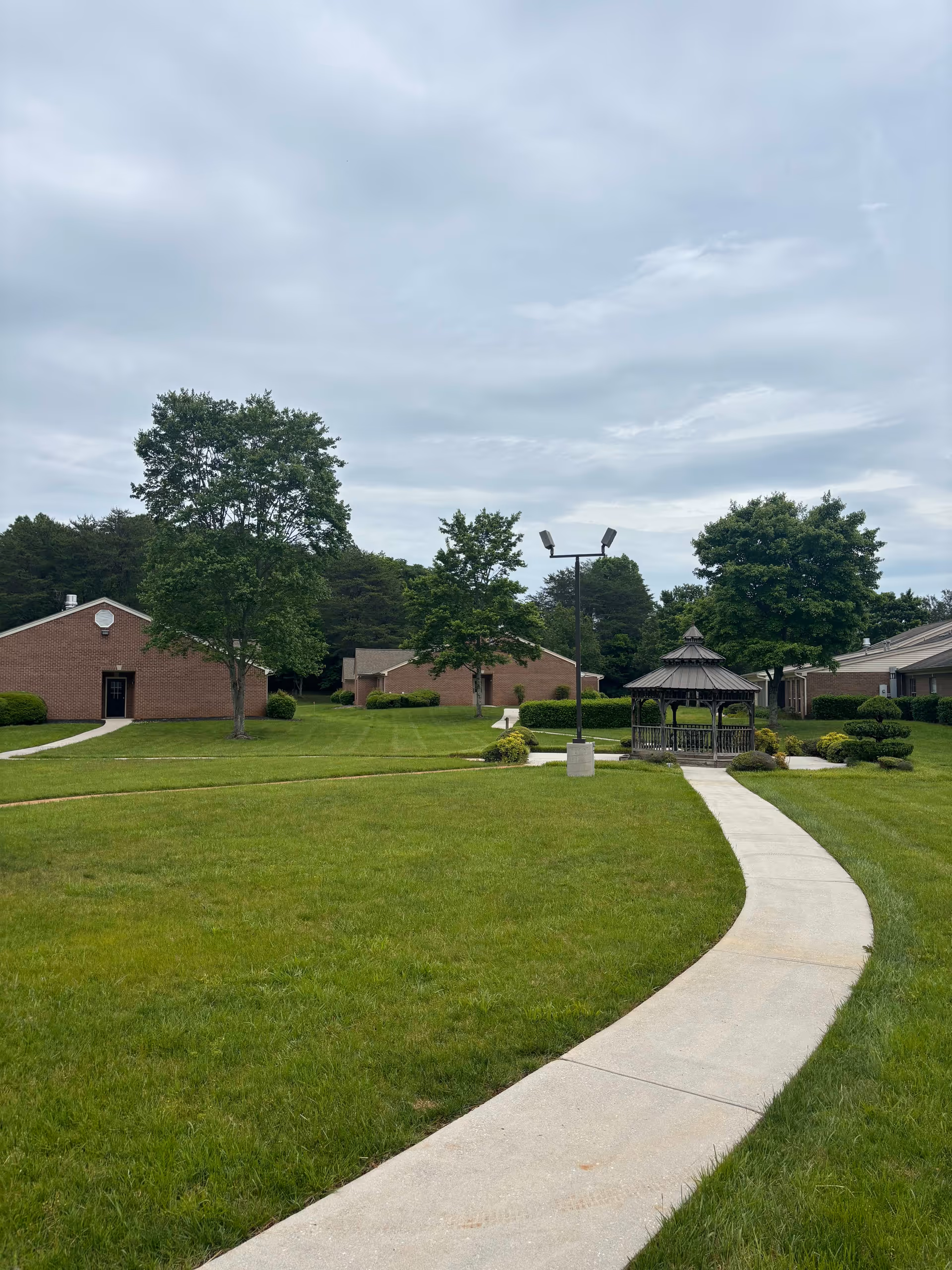 A curved concrete pathway leads through a well-maintained grassy area towards a gazebo surrounded by trees and bushes. Several single-story brick buildings are visible in the background under a cloudy sky.
