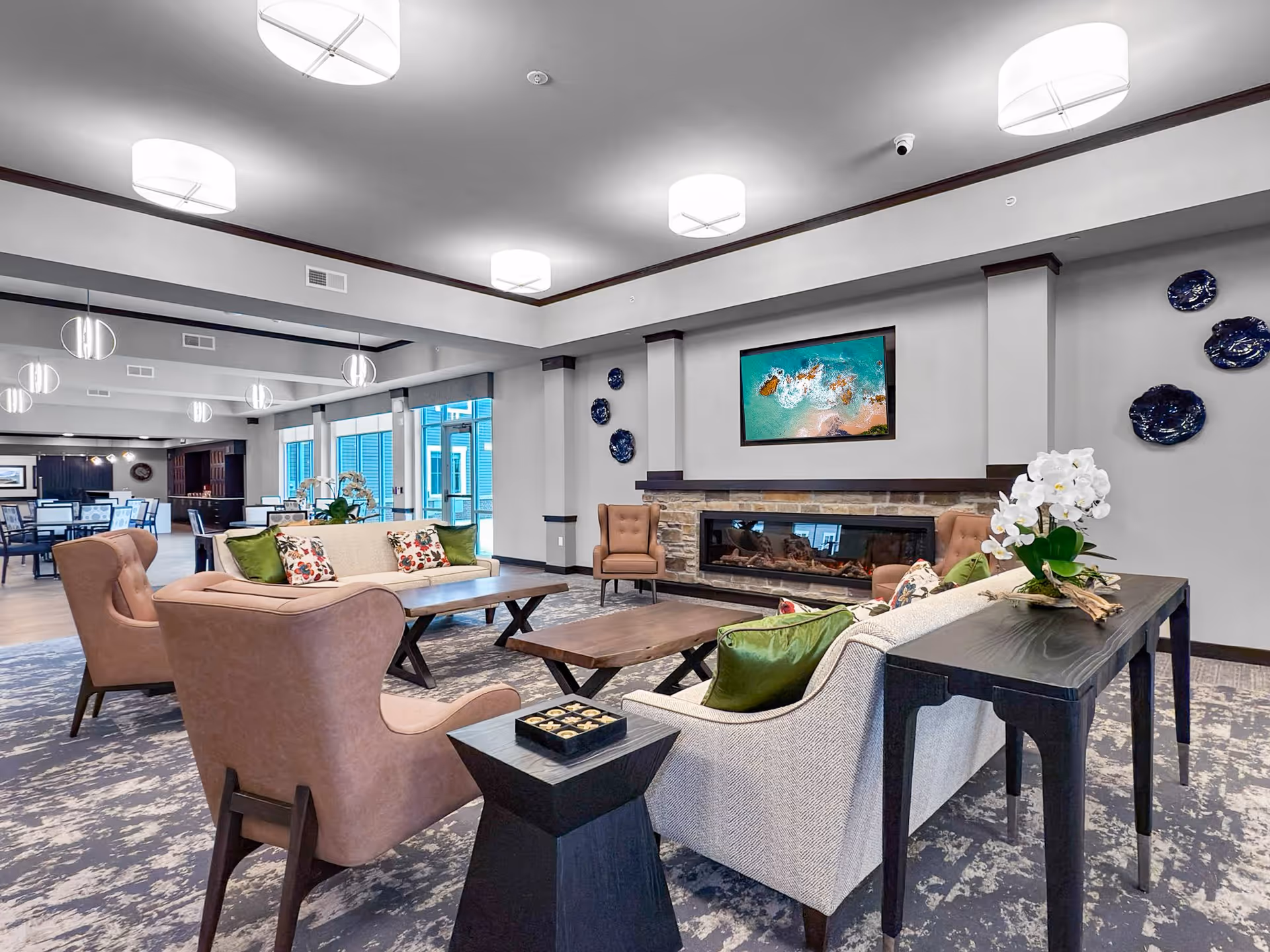 A spacious and modern living room area in a senior living facility featuring comfortable beige and brown armchairs and sofas with green and floral pillows. The room has a large stone fireplace with a flat-screen TV mounted above it, decorative blue wall plates, and a dark wooden table with a white orchid plant. In the background, there is a dining area with tables and chairs, large windows, and contemporary ceiling lights.
