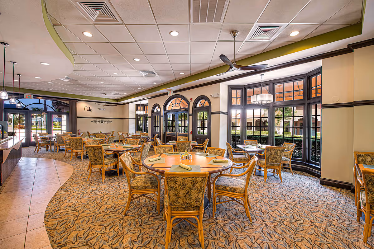 A bright and spacious dining room with multiple round wooden tables surrounded by cushioned chairs with floral upholstery. The room features large windows allowing natural light to fill the space, ceiling fans, and a carpeted floor with a leaf pattern. There is a sign on the wall that reads 'Cafe' and an exit door visible in the background.