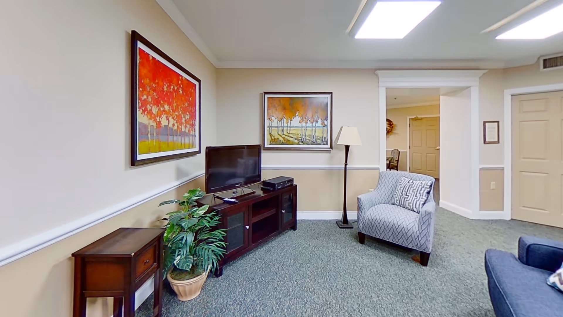 A cozy living room area with a patterned armchair, a dark wooden TV stand holding a flat-screen television and electronic devices, a potted plant, a floor lamp, and two colorful framed paintings of trees on the wall. The room has beige walls with white trim and a green carpeted floor. There is a doorway leading to another room with beige doors.