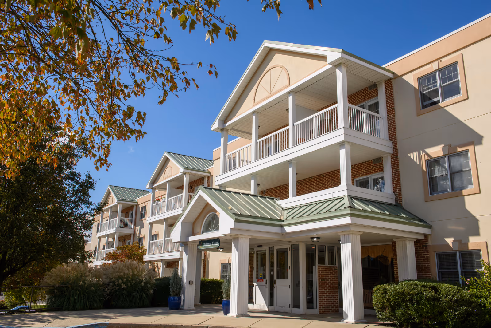 Front entrance and balconies of a three-story senior living building with a covered portico and green metal roofs.