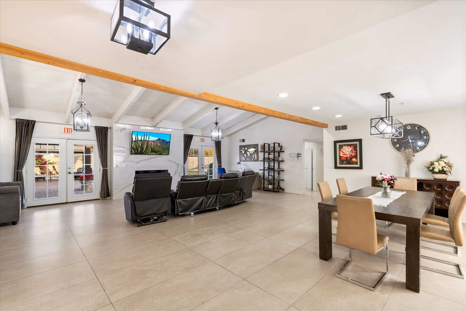 A spacious and bright common area in an assisted living facility featuring a seating area with black recliners facing a wall-mounted TV above a fireplace. The room has high vaulted ceilings with exposed wooden beams and modern hanging light fixtures. To the right, there is a dining area with a dark wooden table and six beige chairs, decorated with flowers and a large wall clock. French doors with dark curtains lead to an outdoor patio area.