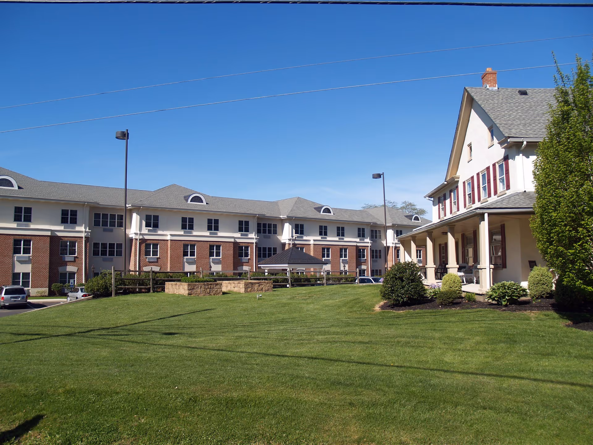 Exterior view of Sacred Heart Senior Living facility showing a large green lawn in the foreground, a multi-story brick and white building in the background, and a smaller white house with red shutters and a porch on the right side under a clear blue sky.