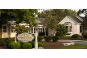 Exterior view of Maple Farm Nursing Center showing a white building with a gabled roof surrounded by trees and shrubs, with a sign in front displaying the facility's name.