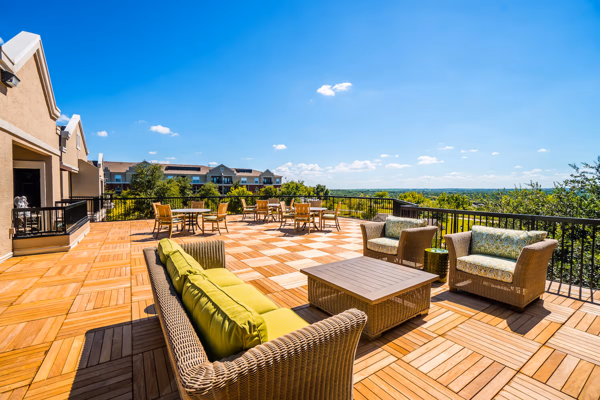 Spacious outdoor patio area with wooden flooring, wicker furniture including a sofa with green cushions, two armchairs with patterned cushions, a coffee table, and several round tables with chairs. The patio is surrounded by a black metal railing and overlooks greenery and distant buildings under a clear blue sky.