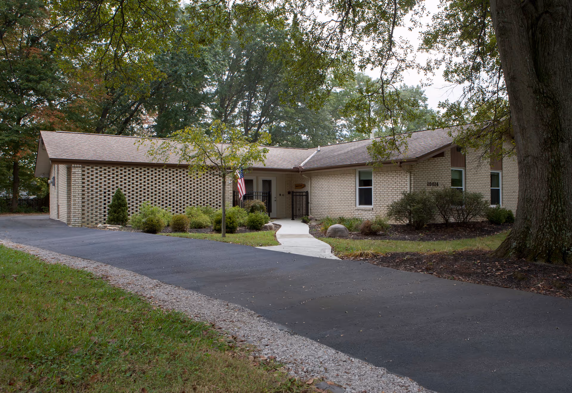 Single-story brick building with a paved driveway and a concrete walkway leading to the entrance. The entrance has a small American flag and a welcome sign above the door. The building is surrounded by trees and shrubs.