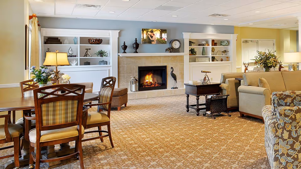 A cozy living room area in a senior living facility featuring a lit fireplace with decorative items on the mantel and built-in shelves on either side. The room has a patterned carpet, a round wooden table with chairs, beige sofas, a floral armchair, and various lamps and plants providing a warm and inviting atmosphere.